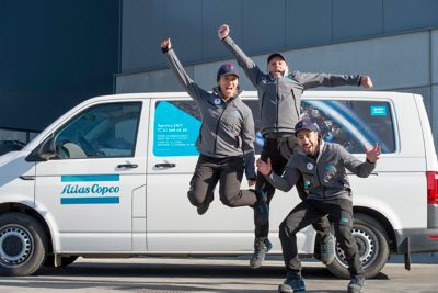 2 hommes et 1 femme sautent de joie devant un véhicule de service Atlas Copco