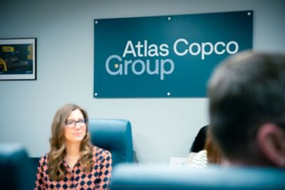 En brunhårig kvinna sitter framför en vägg med en skylt med Atlas Copco Groups logotyp.