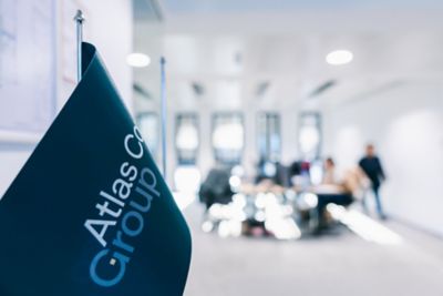 A close-up of Atlas Copco Group flag in focus, while a modern meeting room with people and desks appears in the background
