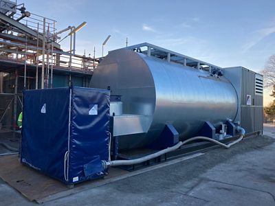 22-ton industrial boiler from Eco Steam and Heating Solutions, outdoors on a concrete pad. The boiler is large, cylindrical, light gray, with pipes and fittings. A smaller box with a blue tarp is in front.