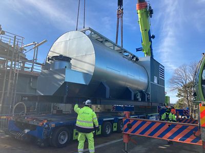 22-ton boiler being lifted by crane. The boiler is large, cylindrical, light gray, with pipes and fittings. Workers in safety gear oversee the operation.