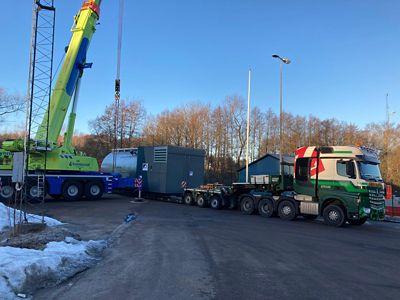Large industrial boiler being transported on a flatbed truck. A crane is lifting the boiler from the truck. Trees and a clear blue sky are in the background.