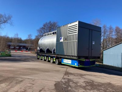 22-ton boiler being transported on a flatbed truck. The boiler is large, cylindrical, light gray, with pipes and fittings. Trees and a clear blue sky are in the background.