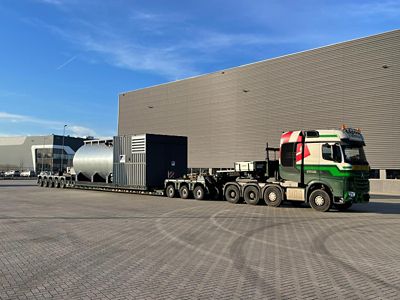 A large, industrial boiler is being transported on a flatbed truck. The truck is parked in a parking lot with a large building in the background.