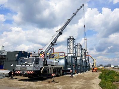 A large crane with a long boom is lifting a chimney of a boiler into place. 