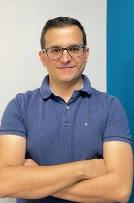 Homem com braços cruzados vestindo camisa polo azul com logotipo bordado, posicionado diante de fundo dividido em branco e azul.