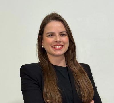 Mulher jovem com cabelos longos e castanhos, vestindo blazer preto com botões dourados e blusa preta. Ela está sorrindo, de braços cruzados, posando em frente a uma parede branca.
