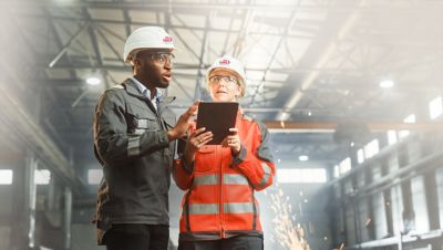 Female industrial engineer wearing a white helmet while standing in a heavy industrial factory behind she talking with workers, Various metal parts of the project