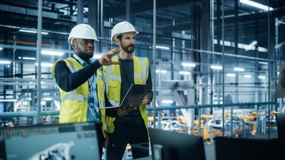 Car Factory: Project Manager and Automotive Engineer in Hard Hat, Talking, Using Laptop. Monitoring, Control, Equipment Production. Automated Robot Arm Assembly Line Manufacturing Electric Vehicles