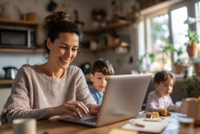 Juggling Hybrid Work and Family Woman Working on Laptop in Kitchen with Kids Playing Nearby