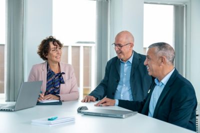 Executives, two older men and one woman, standing around an office table engaged in conversation.