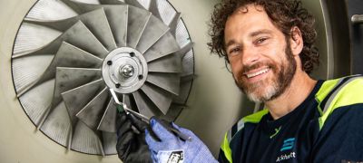 Mann mit Bart und lockigem Haar in Arbeitskleidung und Handschuhen vermisst ein Werkstück und lächelt in die Kamera