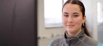 Porträt einer jungen Frau mit zurückgebundenem Haar, grauem Rollkragenpullover und Ohrringen vor unscharfem Hintergrund