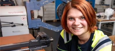Lächelnde Frau mit rotem Haar und Arbeitskleidung in einer Werkstatt mit Maschinen im Hintergrund