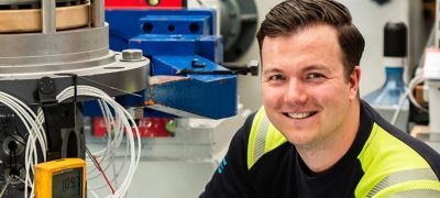Lächelnder Mann in Arbeitskleidung vor blau-grauer Industriemaschine mit Messgerät und Kabeln