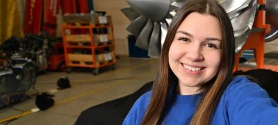 Junge Frau mit langem braunem Haar und blauem Pullover lächelt in einer Werkhalle mit großen Metallteilen im Hintergrund