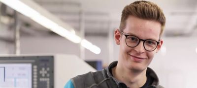 Junger Mann mit Brille und kurzem Haar lächelt in einer Werkhalle mit Maschinen im Hintergrund