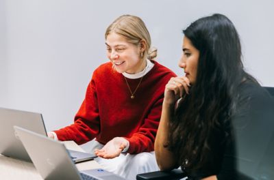 Zwei Frauen sitzen an einem Tisch mit geöffneten Laptops und unterhalten sich