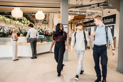 Students conversing while walking in office lobby while others interact at the reception area. A modern office environment with engaging and collaborative atmosphere.