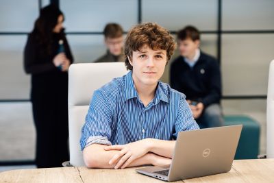Front view of an employee while using a laptop in the office. A blurred background shows other employees engaged in discussion.