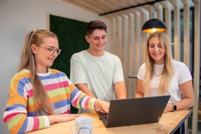 Drei junge Personen stehen an einem Tisch und schauen auf den Laptop
