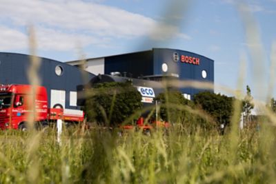A Bosch industrial facility surrounded by greenery, with a red truck and a large black Eco-branded boiler parked outside under a partly cloudy sky.