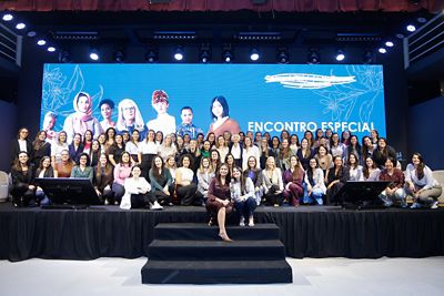 Grupo de pessoas, majoritariamente mulheres, reunido em um palco com fundo azul que exibe a frase 'ENCONTRO ESPECIAL'. Duas telas grandes estão posicionadas nas laterais do palco, e uma mulher está sentada nos degraus à frente do grupo. O fundo também apresenta ilustrações artísticas e imagens de pessoas.