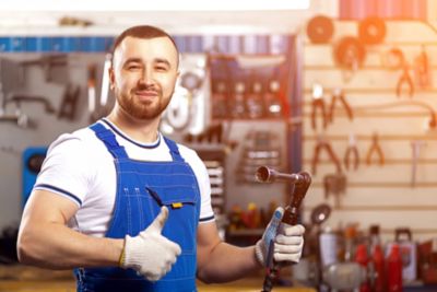 A young man-mechanic in overalls poses at his workplace and holds a pneumatic wrench in his hand, in the background are many tools for repairing a car. Concept Workshop at an automobile repair shop