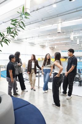 Grupo de seis pessoas em pé em semicírculo dentro de uma instalação industrial, com máquinas ao fundo e um banner azul com a frase "Home of Industrial Ideas". Os indivíduos estão em conversa, com atenção voltada para o centro do grupo. O ambiente é bem iluminado e há uma planta visível à esquerda.