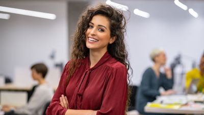 Successful businesswoman standing in creative office and looking at camera. Young latin woman entrepreneur in a coworking space smiling. Portrait of beautiful business woman standing in front of business team at modern agency with copy space.