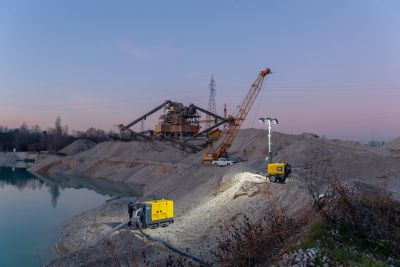 Construction site at dusk beside a calm lake, with cranes, machinery, dirt roads, and a yellow Atlas Copco lighting tower