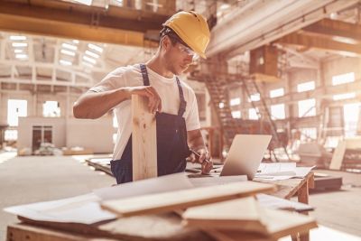 Good-looking young man construction worker standing by the table and holding wooden plank while working on laptop