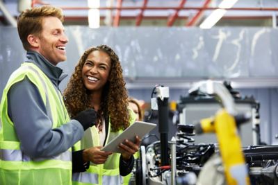 Happy mechanical engineers holding digital tablet by car chassis at factory. Smiling colleagues are standing by car part at automobile industry. They are wearing reflective clothing.