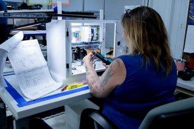 The back of woman sat at a desk assembling a Programmable Logic Controller (PLC) referencing a schemtic that is propped up to her left