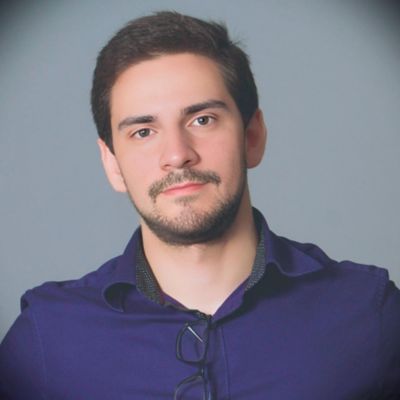 Homem jovem de cabelos castanhos curtos e barba aparada, usando uma camisa social azul escura com óculos pendurados na gola. Ele está posando em frente a um fundo cinza, olhando diretamente para a câmera com expressão neutra.