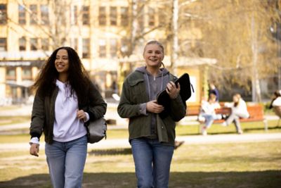 Two young women are smiling while walking across a lawn in a park. One is holding a laptop sleeve, the other carries a backpack over one shoulder. ​