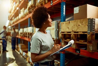 A woman holds a clipboard while checking a box on a warehouse shelf