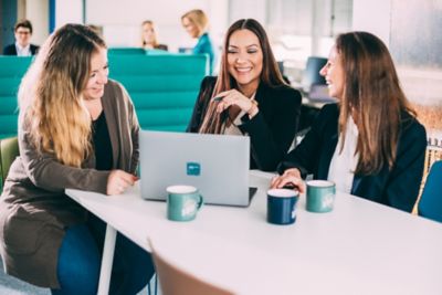 Three women engage in a discussion in office using a laptop. The image illustrates collaboration and teamwork in a modern workspace setting, reflecting productivity and mutual communication.