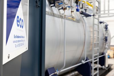 Close-up of the Eco Steam and Heating logo. In the background is a frame-mounted industrial boiler with visible pipes and connections.