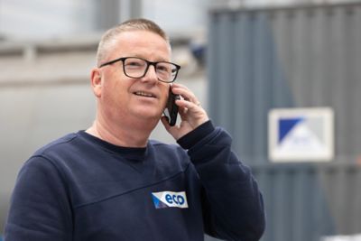A man in a factory setting with a boiler in the background, wearing a sweater with the "Eco" logo, talking on a phone. 