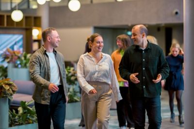 Three employees walk together and talk casually in a workplace corridor with others visible in the background.