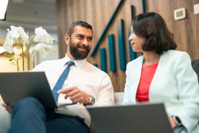 Two colleagues, a man and a woman, sitting down with laptops in their laps in an office reception area.