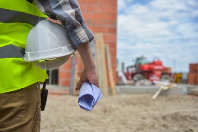Worker holding blueprints and a helmet with background of construction site
