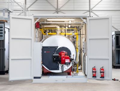 A large industrial boiler in a metal container. The boiler is made of stainless steel and has a variety of pipes and valves attached to it. There are red fire extinguisher next to the container.