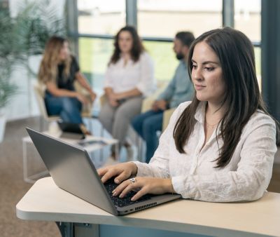 En anställd arbetar med sin laptop i förgrunden, i bakgrunden ser vi en grupp i samtal