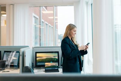 A blonde woman in black blazer standing by the windows in an office checking something on her phone. ​