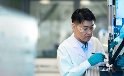 A man with a lab coat, safety goggles and plastic gloves is working on a machine. ​