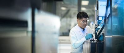 A man with a lab coat, safety goggles and plastic gloves is working on a machine. ​