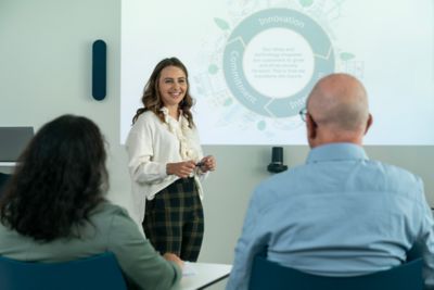 Woman standing in front of a screen, holding a presentation with people in the audience having their backs to the camera. 