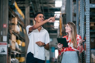 Two employees standing in an aisle between shelves in a warehouse. One is a man pointing something out to the other, a woman holding a tablet. ​